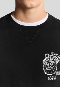 Maglione nero con una stampa grafica bianca di un volto barbuti con un cappello. Il materiale è liscio, con un design a girocollo e dettagli cuciti.