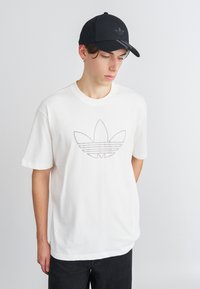 Balts kokvilnas T-krekls ar pelēku izšuvumu Adidas logo, kas attēlo trīs lapu formas. Valkāts ar melnu cepuri, papildināts ar tumšām džinsām.