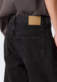 Jeans de denim negro con una etiqueta de cuero marcada "WEEKDAY" en la cintura, presentan bolsillos traseros estándar y un acabado texturizado.
