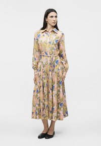 Nő áll, hosszú ujjú, gombos virágmintás ruhát visel, melynek derekán megkötött öv van, fekete lapos cipővel, egyszínű világos háttér előtt.