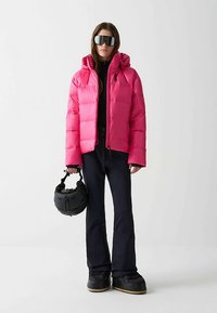 Różowa, ocieplana kurtka narciarska z kapturem, zapinana na zamek, z logo, w połączeniu z czarnymi spodniami i butami. Osoba trzyma czarny kask.