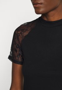 Černé tričko s krátkými krajkovými rukávy. Hladká látka s vysokým výstřihem, krajkové detaily podél ramen a přiléhavý střih.