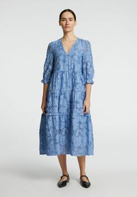 Světle modré maxi šaty s květinovým krajkovým vzorem a tříčtvrtečními rukávy, výstřih do V a volánový design. Outfit doplňují ploché černé boty.