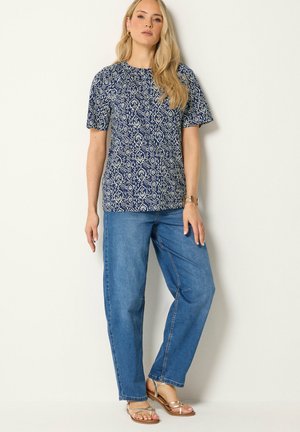 Femme debout contre un mur portant un chemisier bleu à motifs à manches courtes, un jean bleu ample et des sandales beiges.