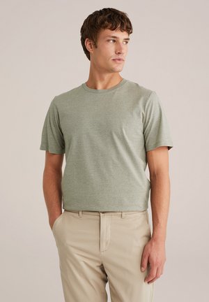 Hombre vestido con una camiseta de cuello redondo verde claro y pantalones beiges, de pie con una mano en el bolsillo, mirando hacia su derecha contra un fondo liso.