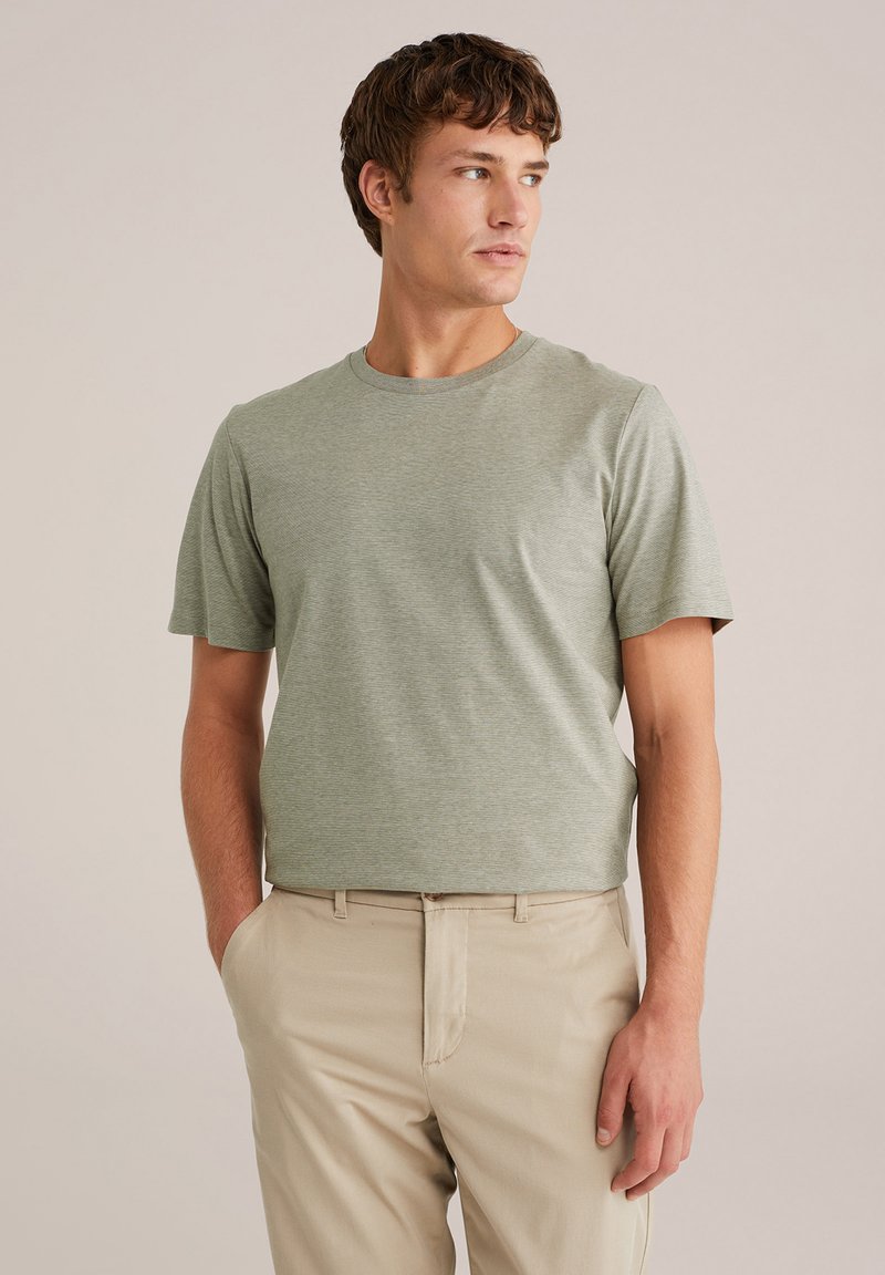 Bărbat îmbrăcat cu un tricou verde deschis și pantaloni bej, stând cu o mână în buzunar, privind spre dreapta pe un fundal simplu.