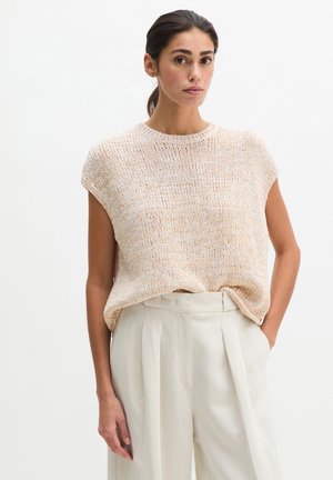 Vrouw met donker haar omhoog gebonden, draagt een mouwloze beige gebreide top en hooggetailleerde crèmekleurige geplooide broek, staand met één hand in de zak.