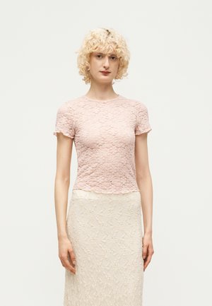 Femme aux cheveux blonds courts et bouclés, portant un top en dentelle rose clair à manches courtes et une jupe en dentelle crème, debout devant un fond uni.