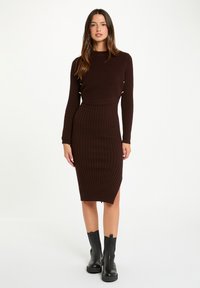 Vestito a maglia marrone a costine con design aderente, maniche lunghe e taglio a lunghezza media. Presenta uno spacco laterale e dettagli decorativi in metallo.