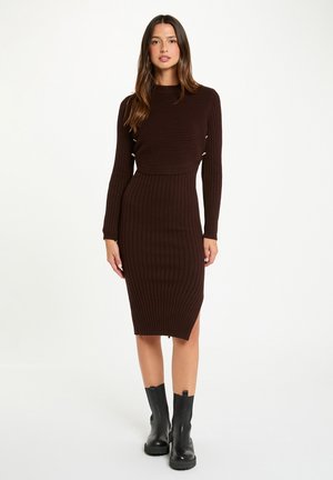 Vestito a maglia marrone a costine con design aderente, maniche lunghe e taglio a lunghezza media. Presenta uno spacco laterale e dettagli decorativi in metallo.