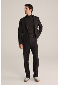 Mand iført mørkebrun jakkesæt med matchende rullekravesweater og hvide sneakers, stående med den ene hånd i lommen mod en ensfarvet baggrund.