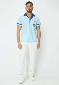Chemise à manches courtes bleu clair avec col marine et rayures, comportant le texte "VIVRE LA VIE" et un accent floral ; assortie à un pantalon blanc texturé.