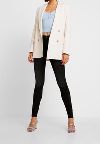 Blazer cruzado crema sobre un top corto de canalé en azul claro, combinado con unos jeans skinny negros y sandalias de tacón azul claro con detalles dorados.