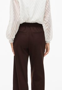 Ruske leve bukser med elastisk talje, der har to baglommer og en glat tekstur, parret med en hvid mønstret bluse.