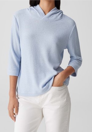 Frau trägt eine hellblaue, strukturierte Kapuzenbluse mit Dreiviertelärmeln und weiße Hose, posiert mit einer Hand in der Tasche.