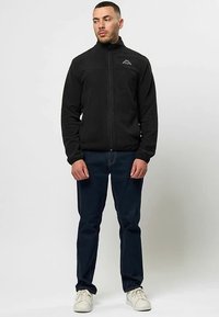 Juodas fliiso švarkas su aukštu apykakle ir užtrauktuku priekyje, ant krūtinės mažas logotipas. Dėvimas su tamsiomis džinsais ir baltais sportbačiais.