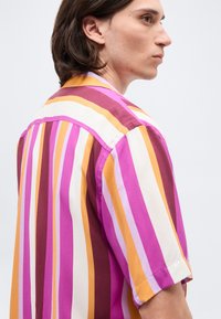 Νεαρός με μεσαίου μήκους καστανά μαλλιά, ντυμένος με κοντομάνικο πουκάμισο με κάθετες ρίγες σε ροζ, κίτρινο, λευκό και μπορντό, κοιτάζοντας δεξιά.