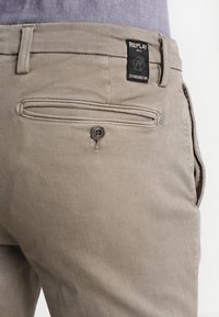 Pantalones de algodón beige con un bolsillo trasero con cierre de botón y una etiqueta negra con texto blanco que muestra detalles de la marca.