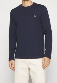 Navy long-sleeve t-shirt made of cotton. Features a crew neckline and a small green logo on the chest. Styled with light-colored pants.