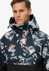 Zwart en meerkleurige windjack met abstracte patronen, rits bij de hals, verstelbare capuchon en "SUPERDRY" logo aan de voorkant.