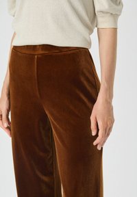 Pantaloni din catifea maro cu o textură netedă, design cu picioare largi și talie plată vizibilă în partea de sus, completate de un top de o culoare deschisă.