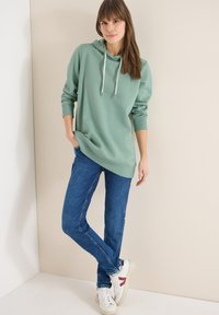 Femme en sweat à capuche vert, jean bleu et baskets blanches, debout de manière détendue contre un mur beige, une main dans la poche et les jambes croisées.