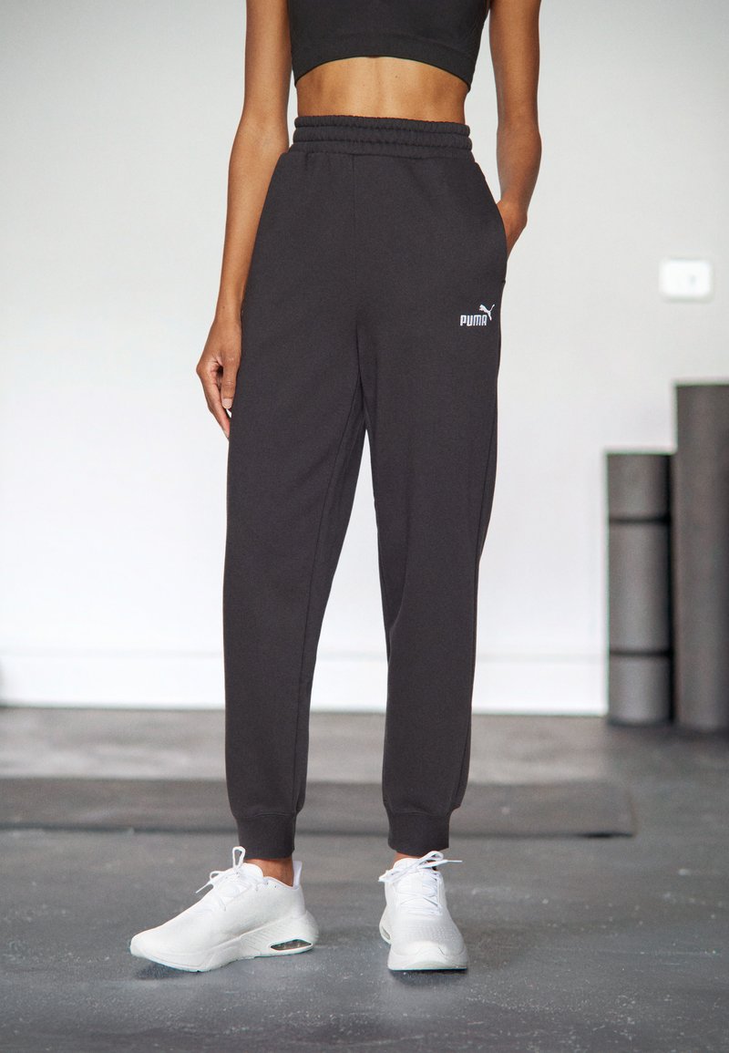 Pantaloni della tuta neri con elastico in vita, con il logo Puma bianco. Abbinati a scarpe da ginnastica bianche su un pavimento grigio.