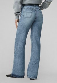 Asmuo, dėvintis šviesiai mėlynas plačias džinsines kelnes ir pilką džemperį, stovintis su viena ranka ant klubų.