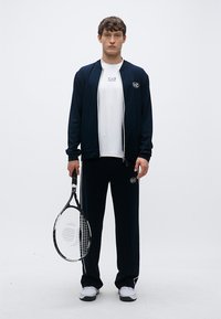 Pánske športové oblečenie zahŕňa tmavomodrú mikinu na zips, biele tričko, tmavomodré nohavice s logom a drží tenisovú raketu.