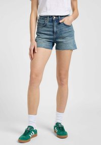 Szorty dżinsowe w jasnoniebieskim kolorze z postrzępionym dołem, wysoka talia i dwie przednie kieszenie, noszone z zielonymi trampkami i białymi skarpetkami.