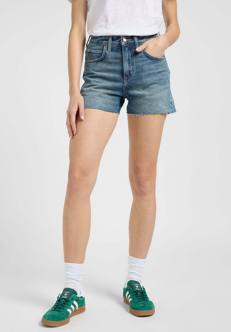 Szorty dżinsowe w jasnoniebieskim kolorze z postrzępionym dołem, wysoka talia i dwie przednie kieszenie, noszone z zielonymi trampkami i białymi skarpetkami.
