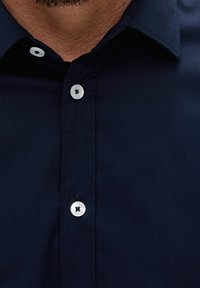 Námořnická modrá košile na knoflíky s hladkou texturou, s bílými knoflíky a konfekčním límcem. Viditelné prošívání podél légy.
