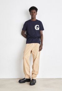 Camiseta marina con un gran logo blanco "G" en el pecho, combinada con pantalones cargo beiges y zapatos de plataforma negros. Texturas de tela suaves y casuales.