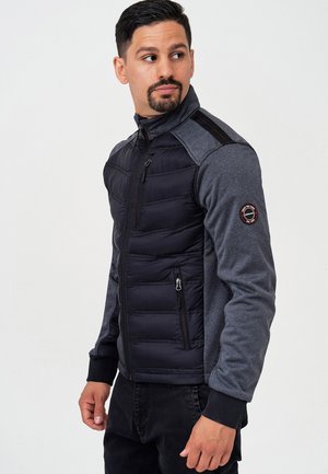 Sort og grå jakke med en puffy front, lynlåslommer, ribbede manchetter og en høj krave. Har et logo på skulderen.
