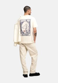 Camiseta de algodón en color blanco roto con un estampado gráfico azul en la parte posterior, combinada con pantalones en color crema y calzado negro.