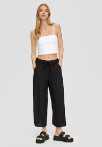 Witte cropped tanktop met dunne bandjes, gecombineerd met zwarte wijde broek met een elastische tailleband en trekkoord. Zwarte platform sandalen.