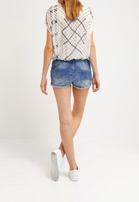 Dámský outfit sestávající z volného béžového topu s černým geometrickým vzorem a modrých džínových kraťasů. Doplněno bílými teniskami.