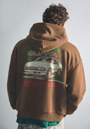 Personne portant un sweat à capuche marron avec un graphisme de voiture Toyota Celica Supra blanc et un logo Toyota rouge dans le dos, faisant face à l'opposé de la caméra.