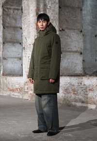 Homme portant un parka à capuche vert olive et un jean large bleu, debout à l'intérieur devant un mur de briques texturé.