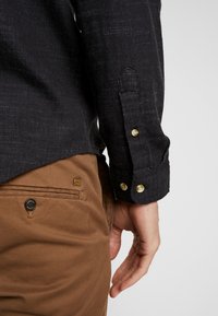 Musta teksturoitu paita, jossa nappikiinnitteiset hihansuut, yhdistettynä ruskeisiin housuihin, joissa on laastaritasku ja musta nappi, esitellen kontrastoivia värejä ja materiaaleja.