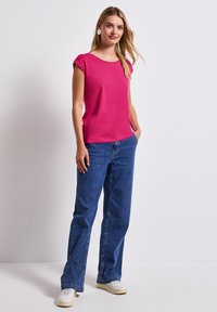 Fuchsiový top s krátkým rukávem a kulatým výstřihem, v kombinaci s volnými modrými džíny a bílými teniskami. Model stojí s rukama v kapsách.