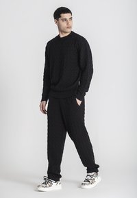 Suéter negro de punto con un patrón de cable texturizado, combinado con pantalones negros a juego. Zapatillas blancas con acentos metálicos y texto.