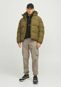 Mann mit schwarzer Kappe, olivgrünem Steppjacke, schwarzem Hoodie, beigen Cargohosen und schwarzen sowie grauen Sneakern, der vor einem schlichten Hintergrund steht.