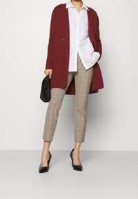 Burgunderfärgad lång cardigan över en vit skjorta, ihop med beige rutiga tapered byxor och svarta högklackade skor. Svart clutch hålls i handen.