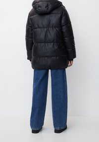 Doudoune noire à capuche, avec des sections matelassées et une longueur mi-cuisse, associée à un jean bleu à jambes larges et des chaussures noires.