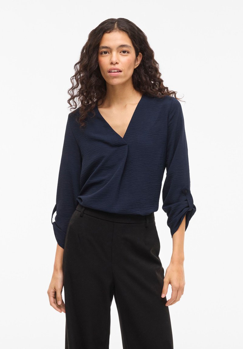 Μπλούζα σε ναυτικό μπλε με V-λαιμόκοψη και υφή υφάσματος, με μακριά μανίκια που είναι γυρισμένα στις μανσέτες. Συνδυασμένη με μαύρο παντελόνι φαρδύ στο πόδι.
