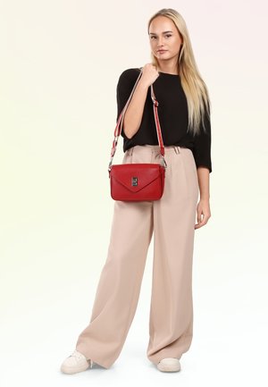 Jeune femme aux longs cheveux blonds portant un haut noir, un pantalon beige large, des baskets blanches, tenant un sac bandoulière en cuir rouge.