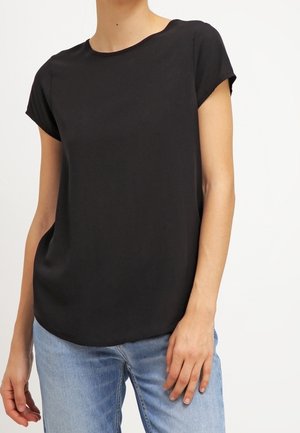 Vrouw draagt een effen zwart topje met korte mouwen, lichtjes in een lichtblauwe spijkerbroek gestopt, tegen een witte achtergrond.