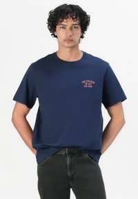Camiseta de algodón azul marino de mangas cortas y cuello redondo. Presenta un pequeño texto rojo "PARA EL VIAJE DE LA VIDA EST. 1947" en el pecho.