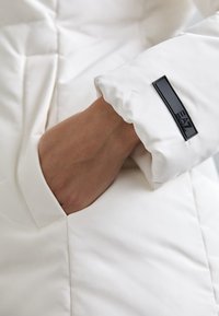 Ruka unutar džepa bijele podstavljene jakne s crnom oznakom EA7 Emporio Armani na rukavu.